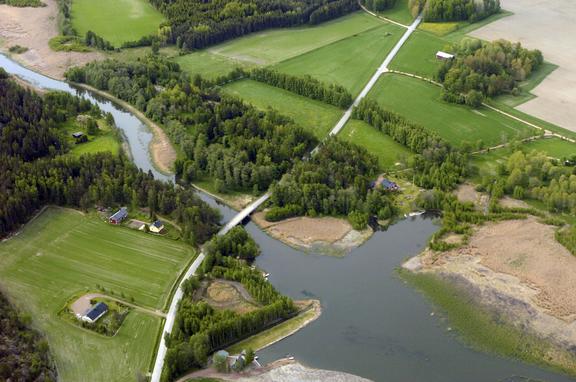 Aerial photo of a rural landscape An aerial view of a rural landscape where a winding river or narrow waterway flows through fields and wooded areas. A road crosses the water via a bridge. The image also shows scattered houses and farm buildings, extensive green farmland, and reed-covered shoreline areas along the water.